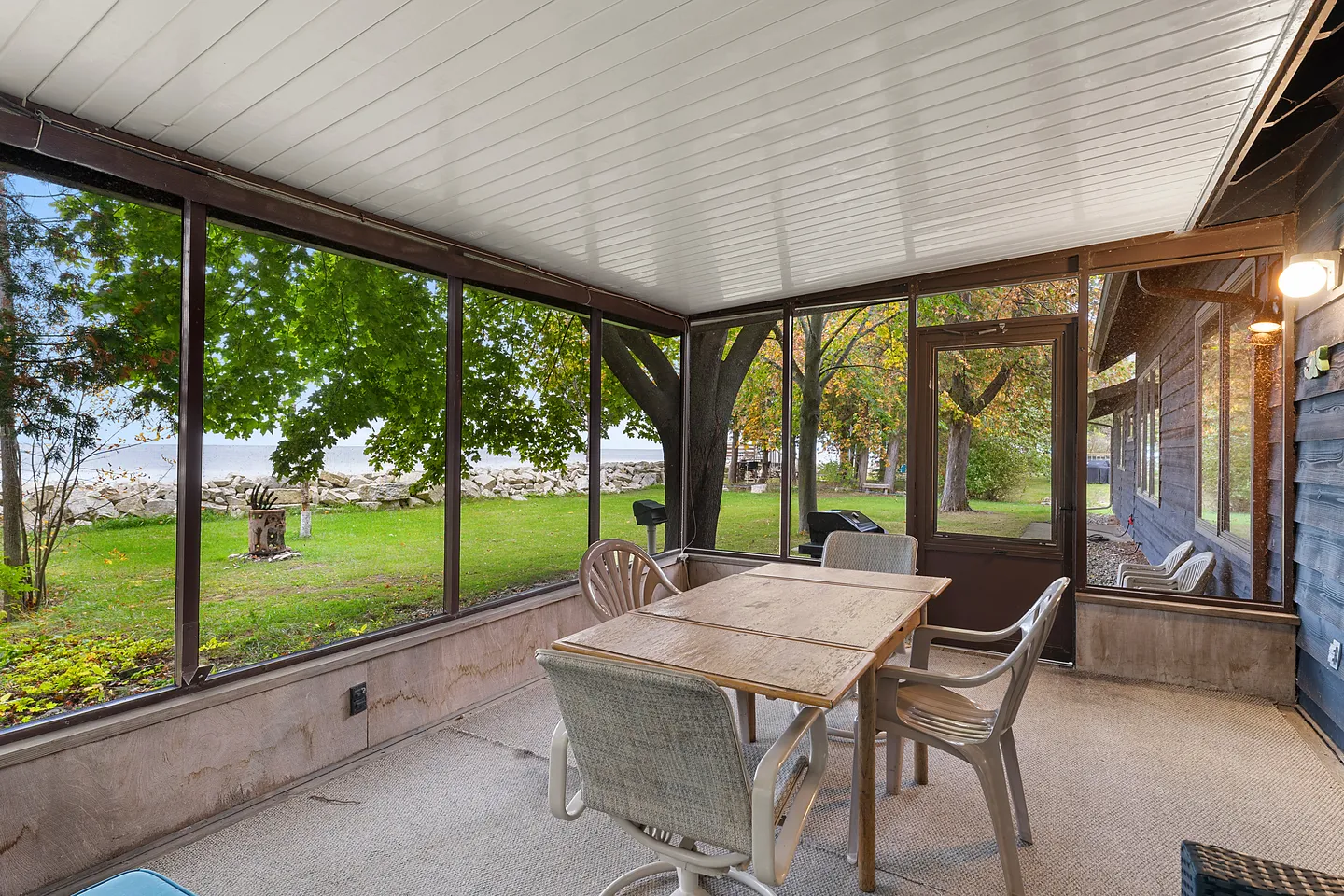 Screened porch furnished with a dining table and chairs, offering views of the grassy yard and water.