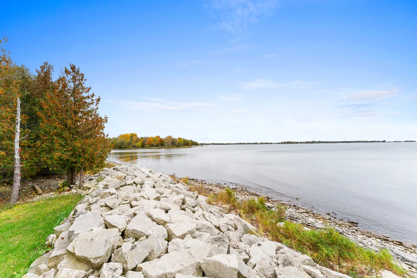 A rock-lined shoreline along a wide body of water with trees showing early autumn foliage under a blue sky.