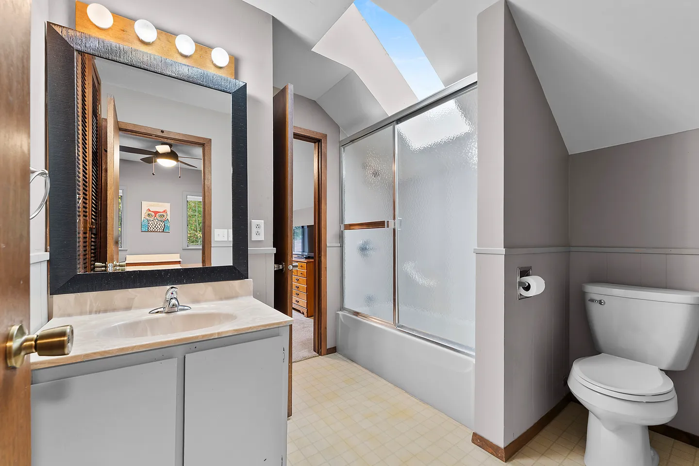 Bathroom with a vanity and mirror, bathtub-shower combination with sliding frosted glass doors, toilet, and a skylight.