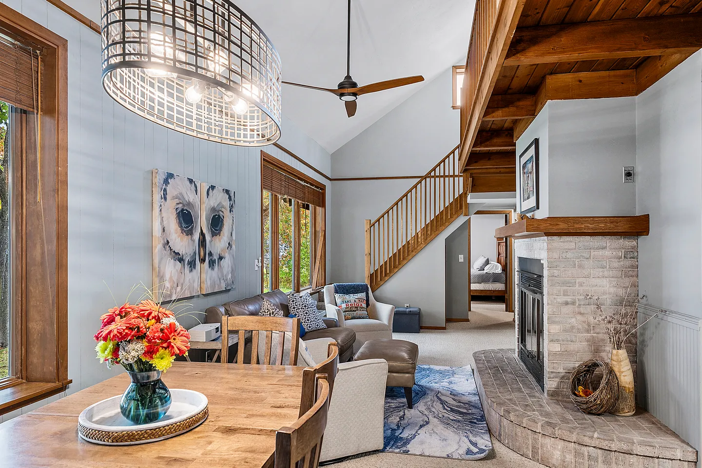 Open living and dining area featuring a brick fireplace, vaulted ceilings, wooden staircase, and dining table with overhead chandelier.