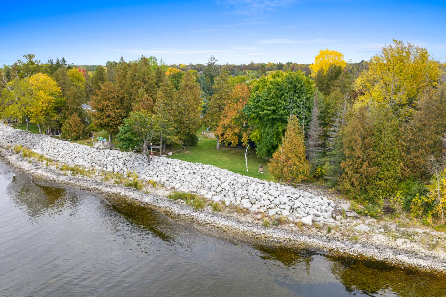 Elevated view of a rocky shoreline with riprap barrier fronting a grassy yard and autumn trees next to water.