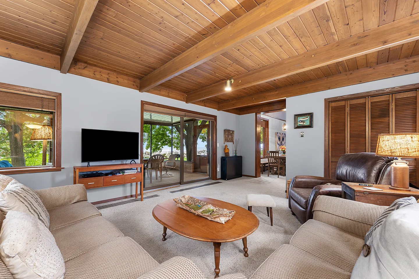 Living room with wood-paneled ceiling, exposed beams, upholstered sofas, an oval coffee table, a flat-screen TV, and sliding glass doors leading outside.