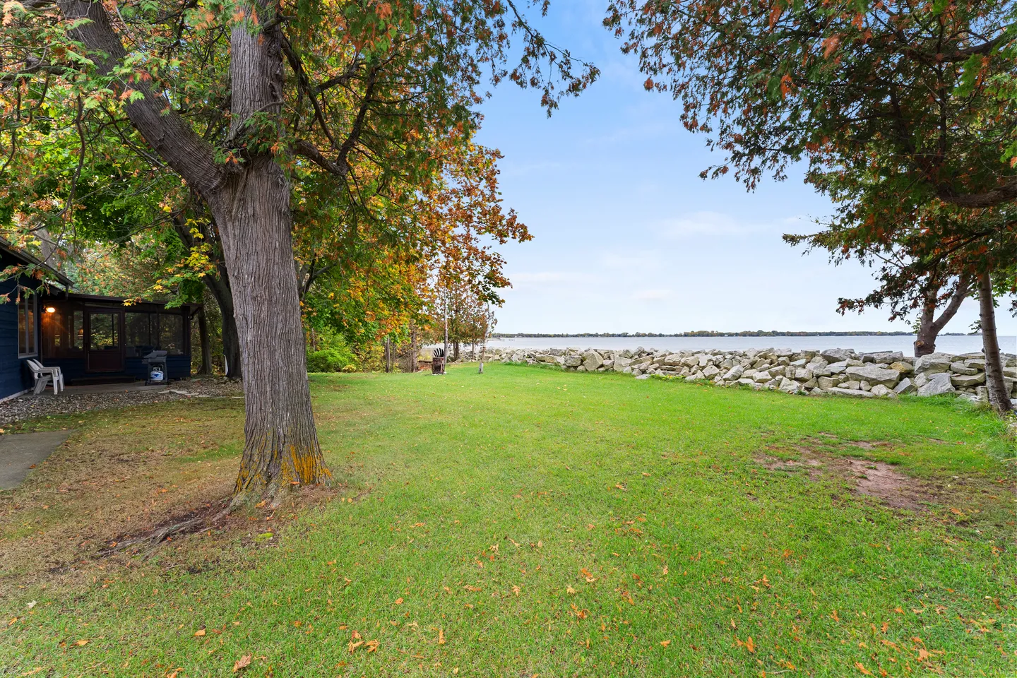 A wide, grassy backyard framed by mature trees, leading toward a stone retaining wall along the edge of a lake.