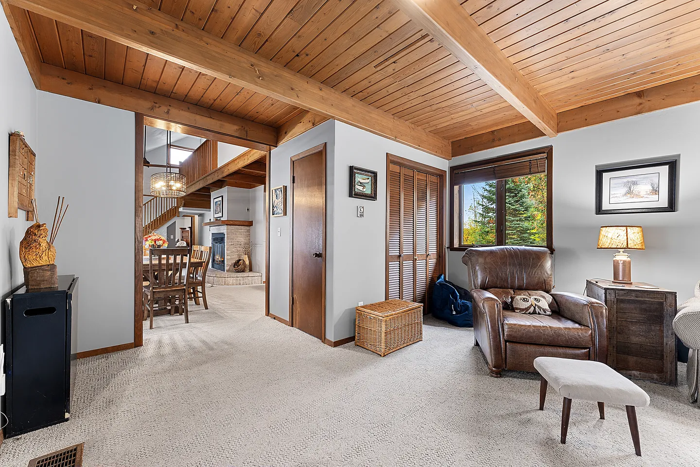 Carpeted sitting area with a wood-paneled ceiling, leather armchair, footstool, and an open doorway leading to a dining area and stone fireplace.