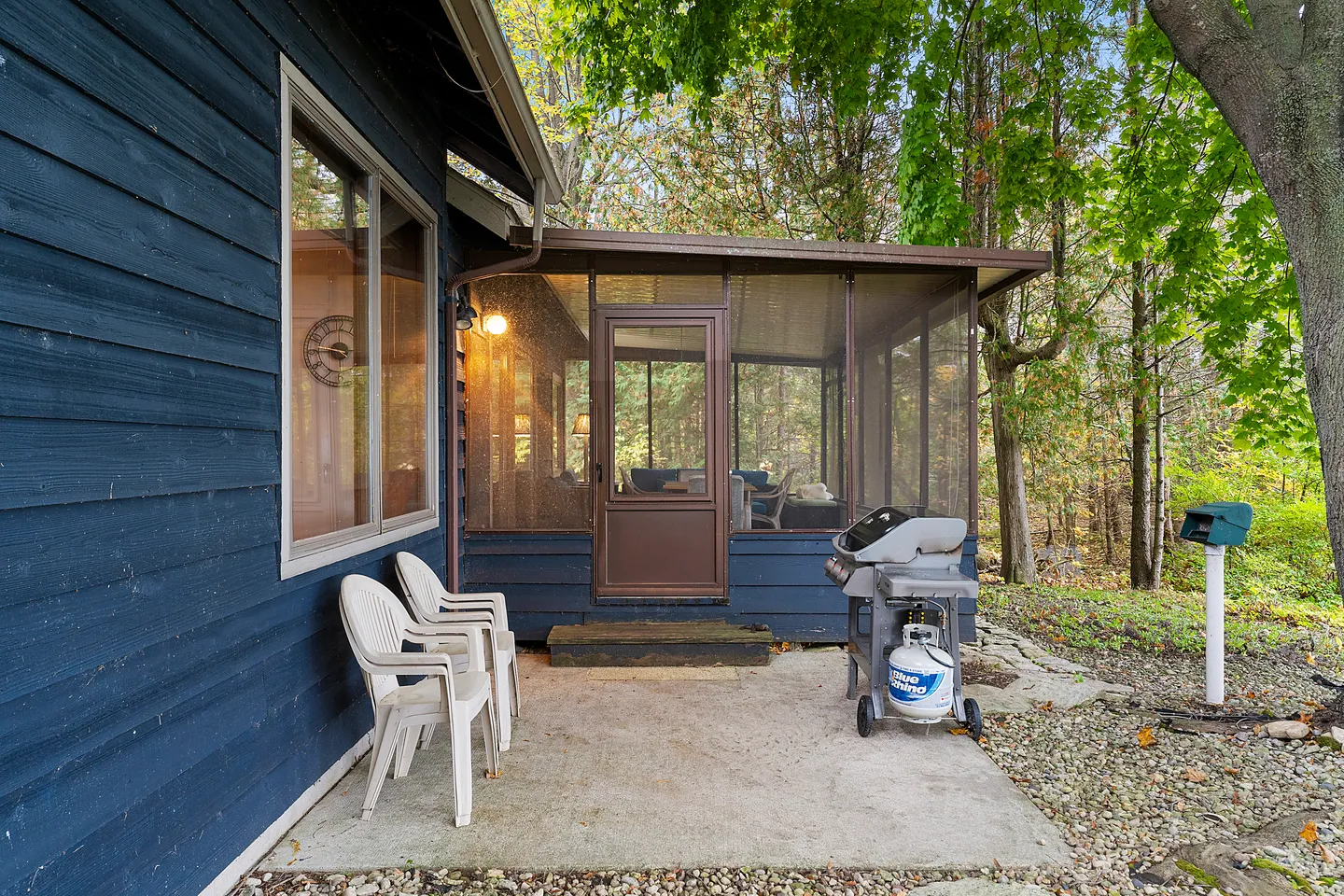 Concrete patio with plastic chairs, a gas grill, and an entrance to a screened porch outside a blue-sided house.