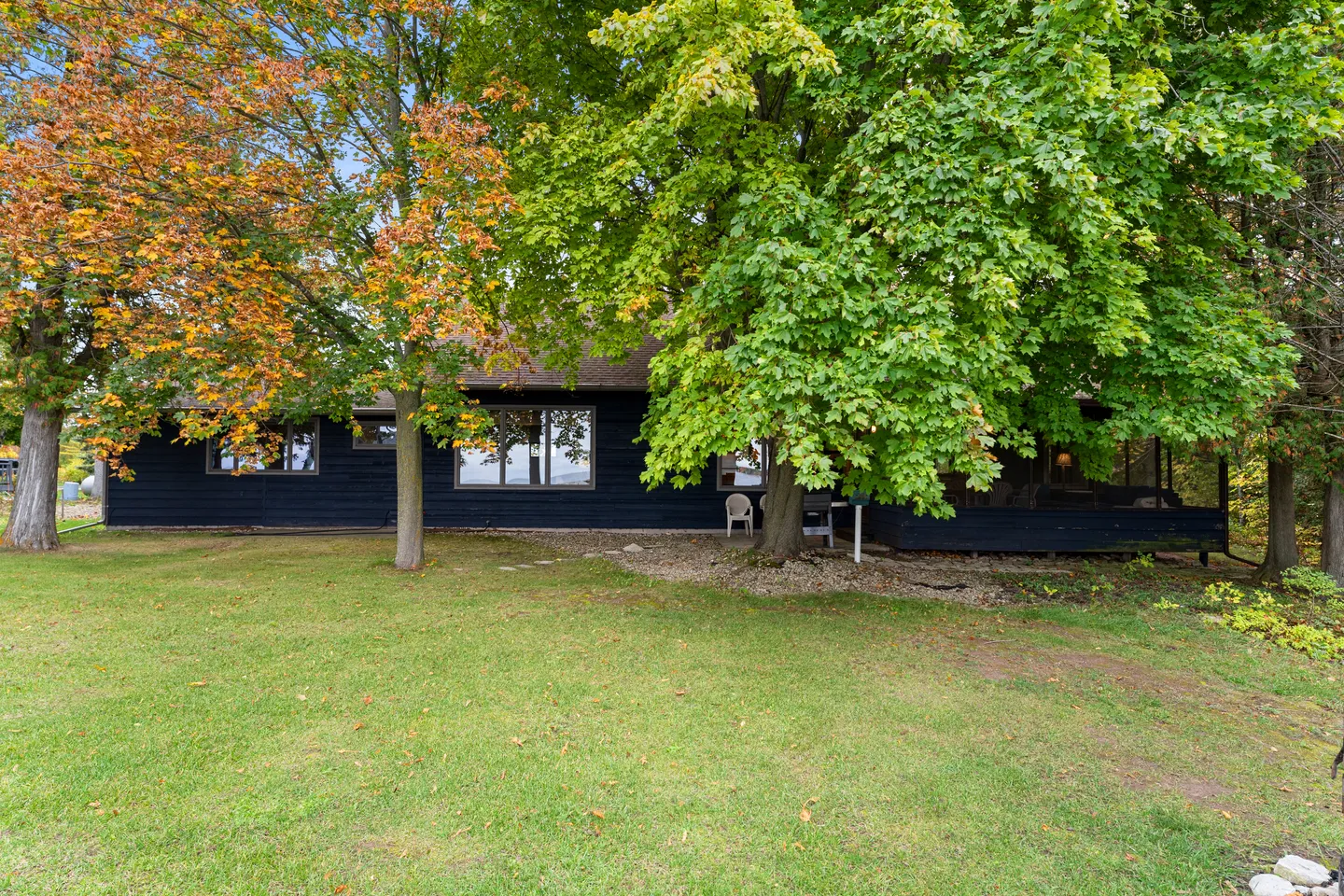 Exterior of a single-story dark house partially shaded by large leafy trees on a grassy lawn.