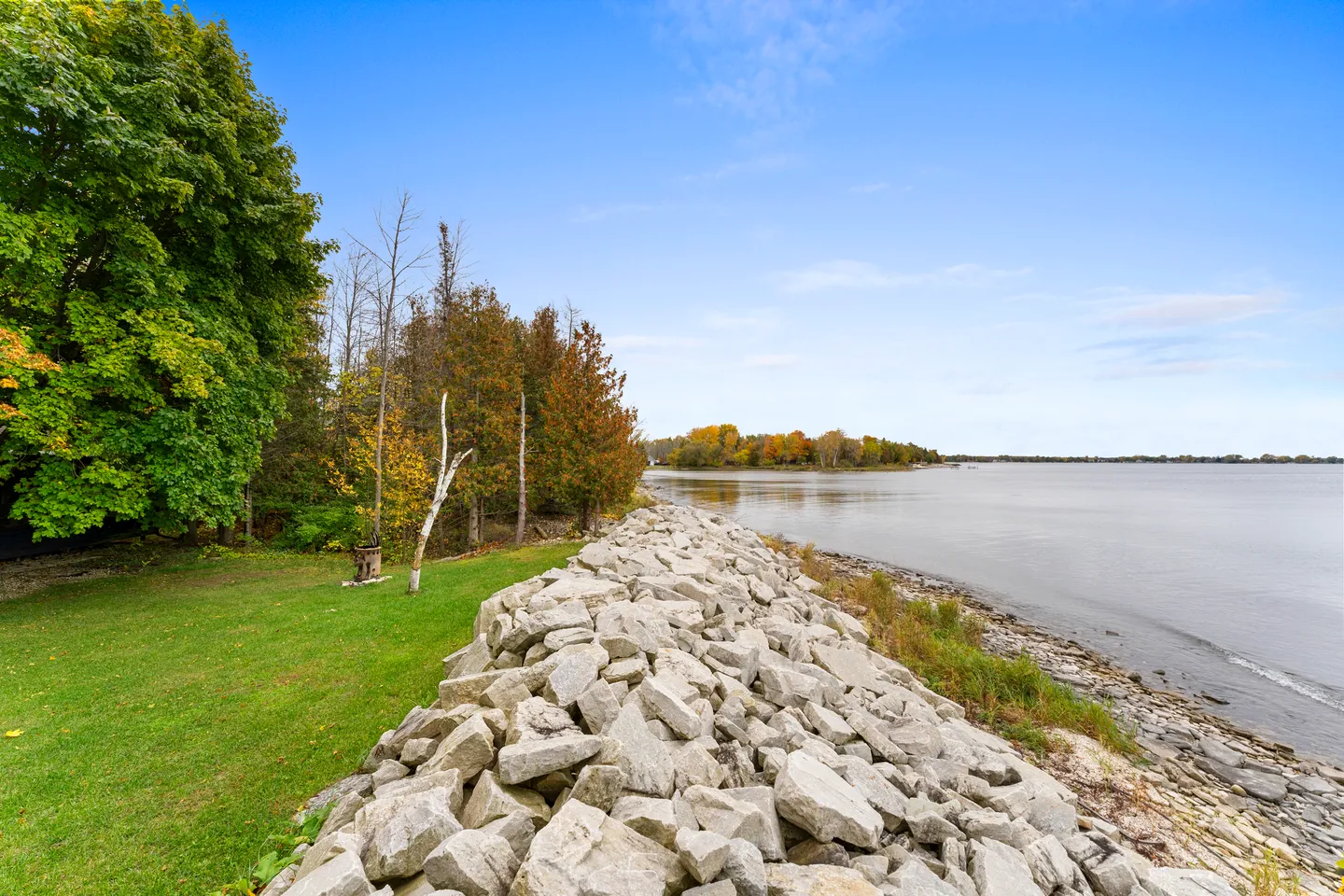 A rocky breakwater shoreline next to a grassy lawn bordering a wide lake under a blue sky.