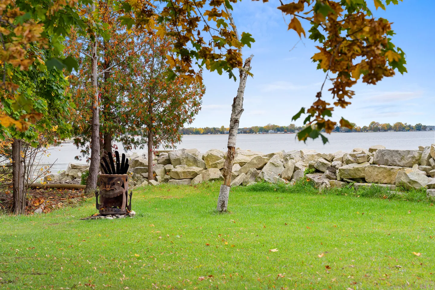 A grassy lawn by a rocky shoreline overlooking an open body of water with a metal fire pit styled like a tiki head.