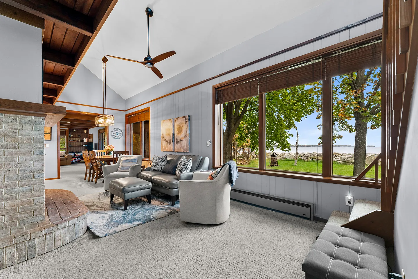 Living room with vaulted ceiling, brick fireplace hearth, leather couch, and large windows overlooking a waterfront lawn.
