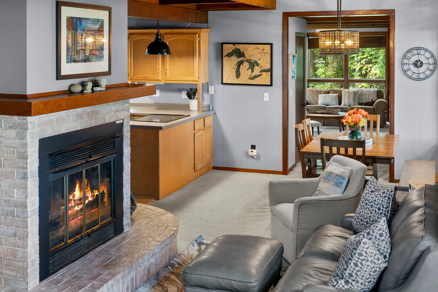 Open-concept living room featuring a lit brick fireplace, cozy seating, and a view into the dining area and kitchen.