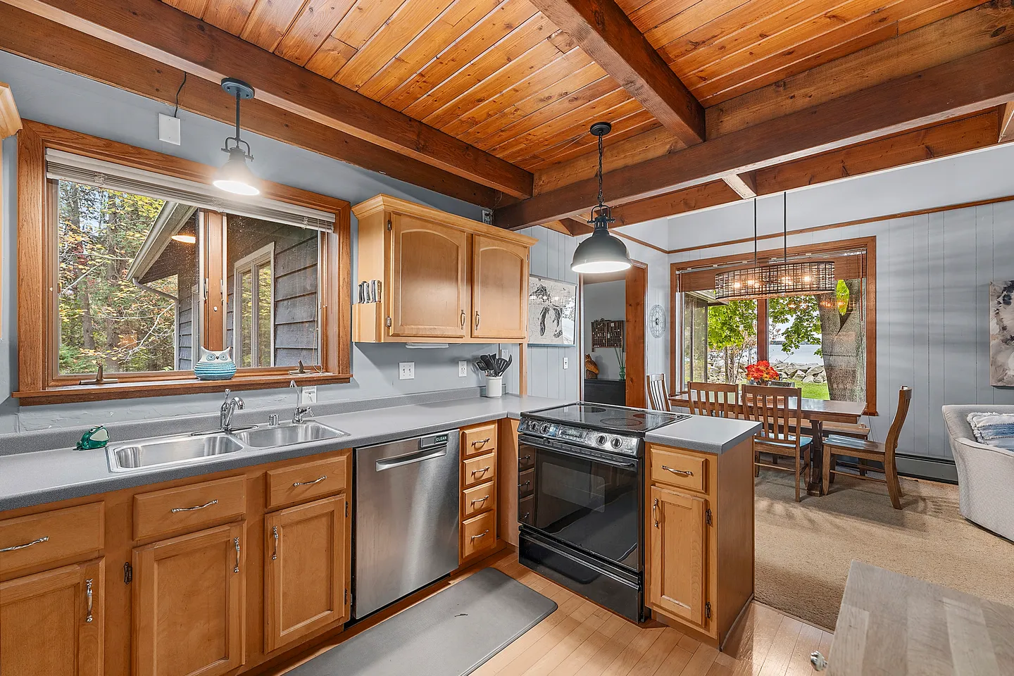 Kitchen with wooden cabinetry, stainless steel dishwasher, dark stove, and wood-plank ceiling leading into an adjacent dining and living space.
