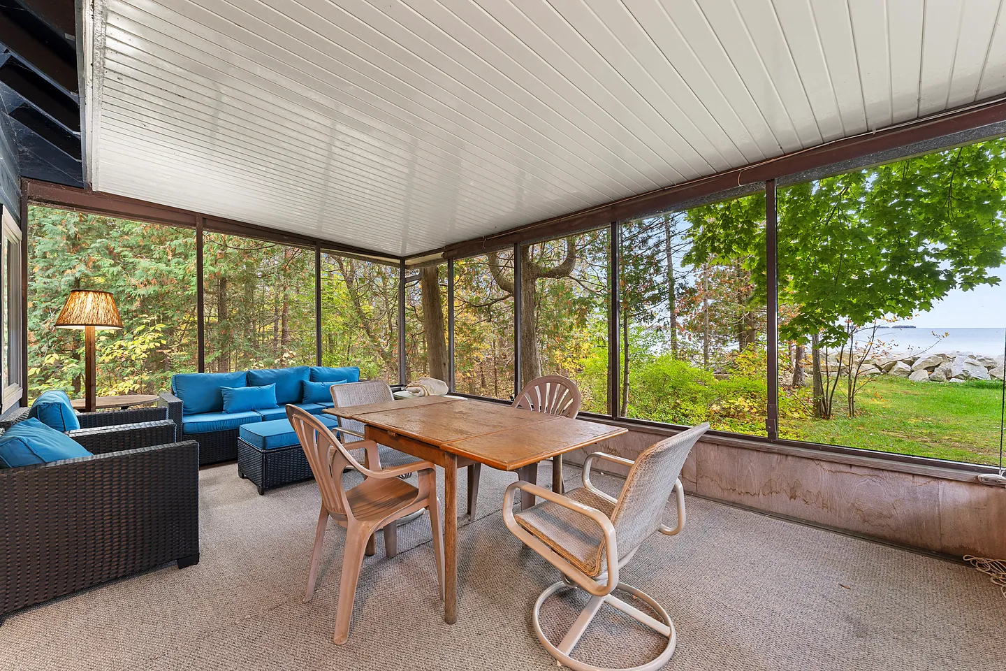 Screened-in porch furnished with a dining table, chairs, and outdoor sofa overlooking a tree-lined shore and water.