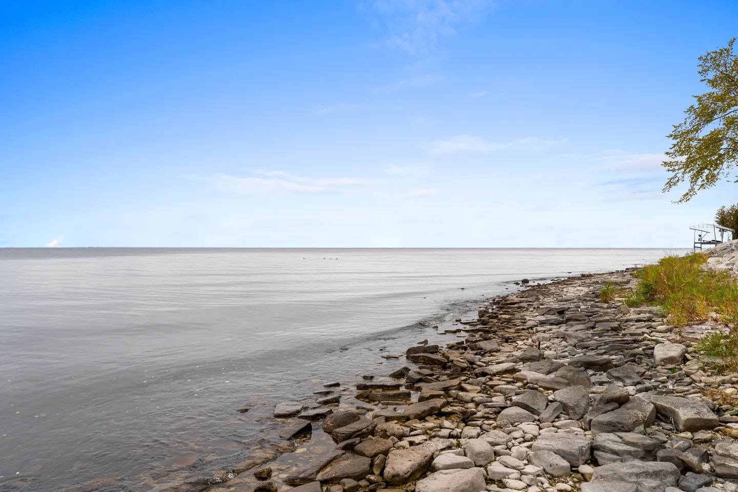 A wide view of a calm, expansive lake shoreline lined with flat rocks and stones under a bright blue sky.