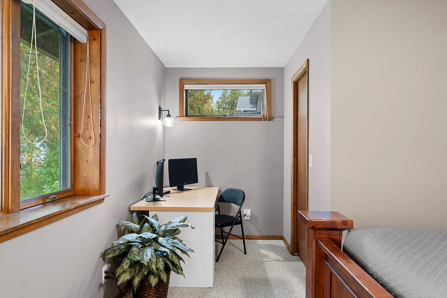 A dedicated home office nook with a desk, two computer monitors, a folding chair, windows, and a potted plant.