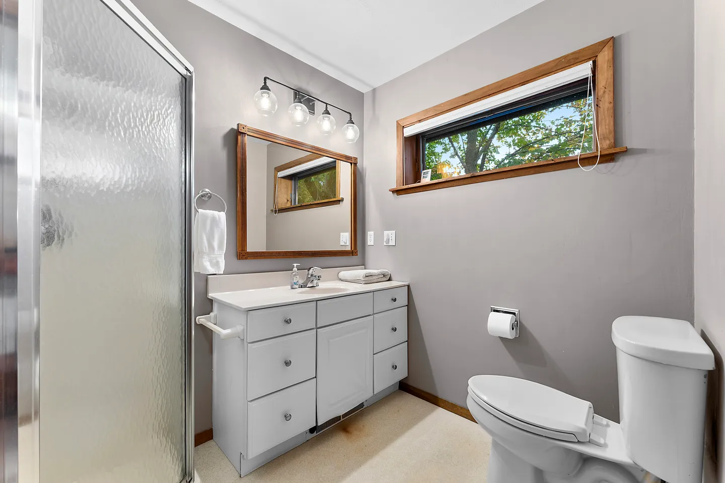 Bathroom with a glass-enclosed shower, a white vanity with drawers, a wood-framed mirror, and a toilet next to a clerestory window.