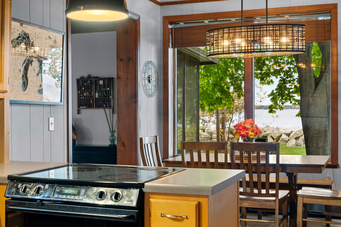 Kitchen counter and stove in the foreground looking toward a dining table with wooden chairs near large windows overlooking a lake.