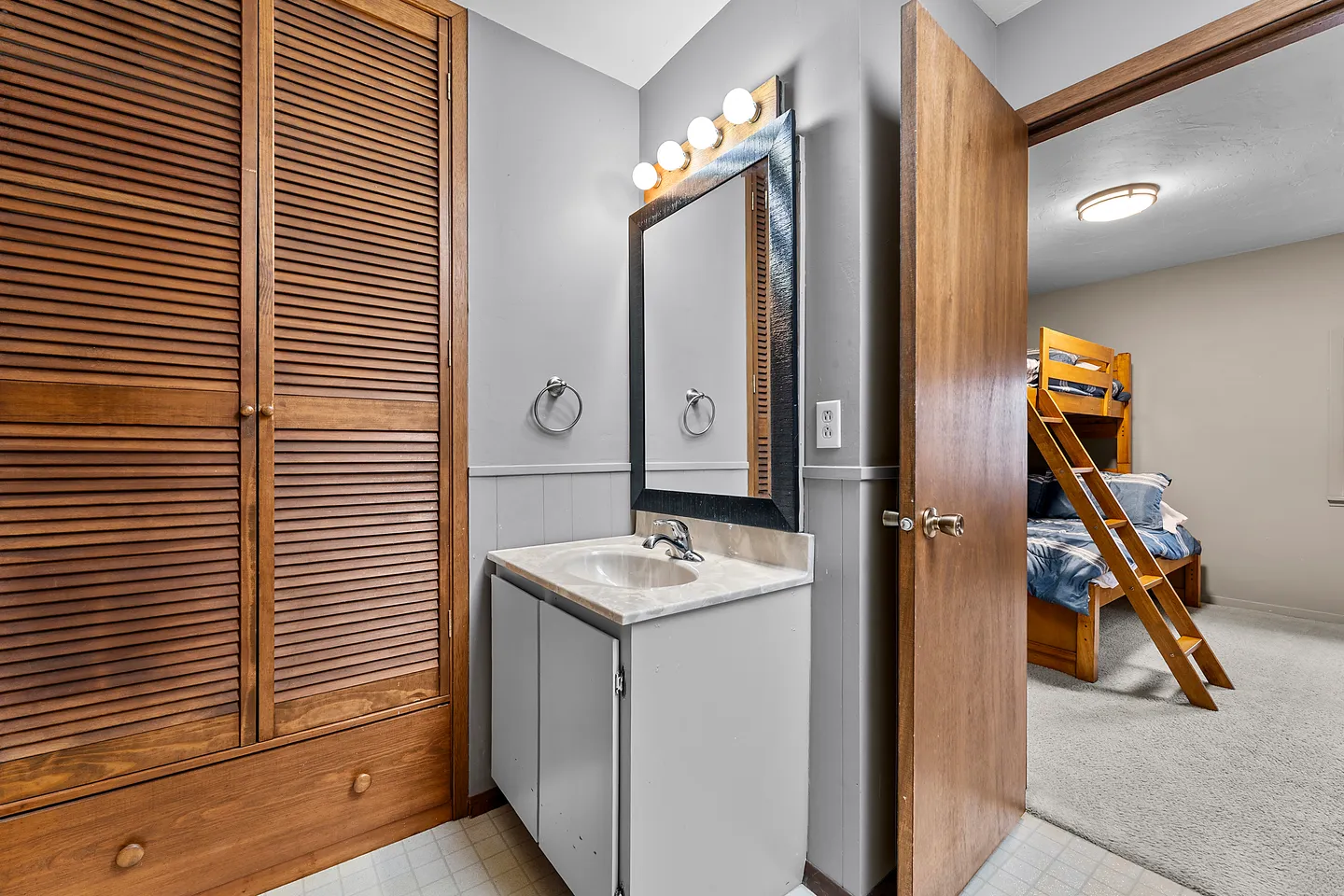 Bathroom vanity with framed mirror and louvered wooden closet doors, with a view into an adjacent bedroom with bunk beds.