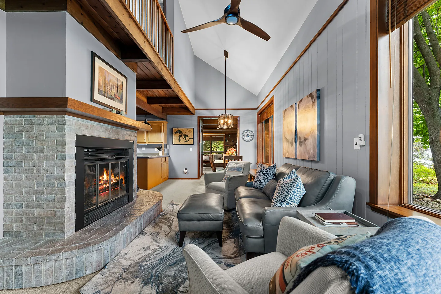 Living room with high vaulted ceilings, a brick-front fireplace, leather seating, and views into an adjoining kitchen and dining area.