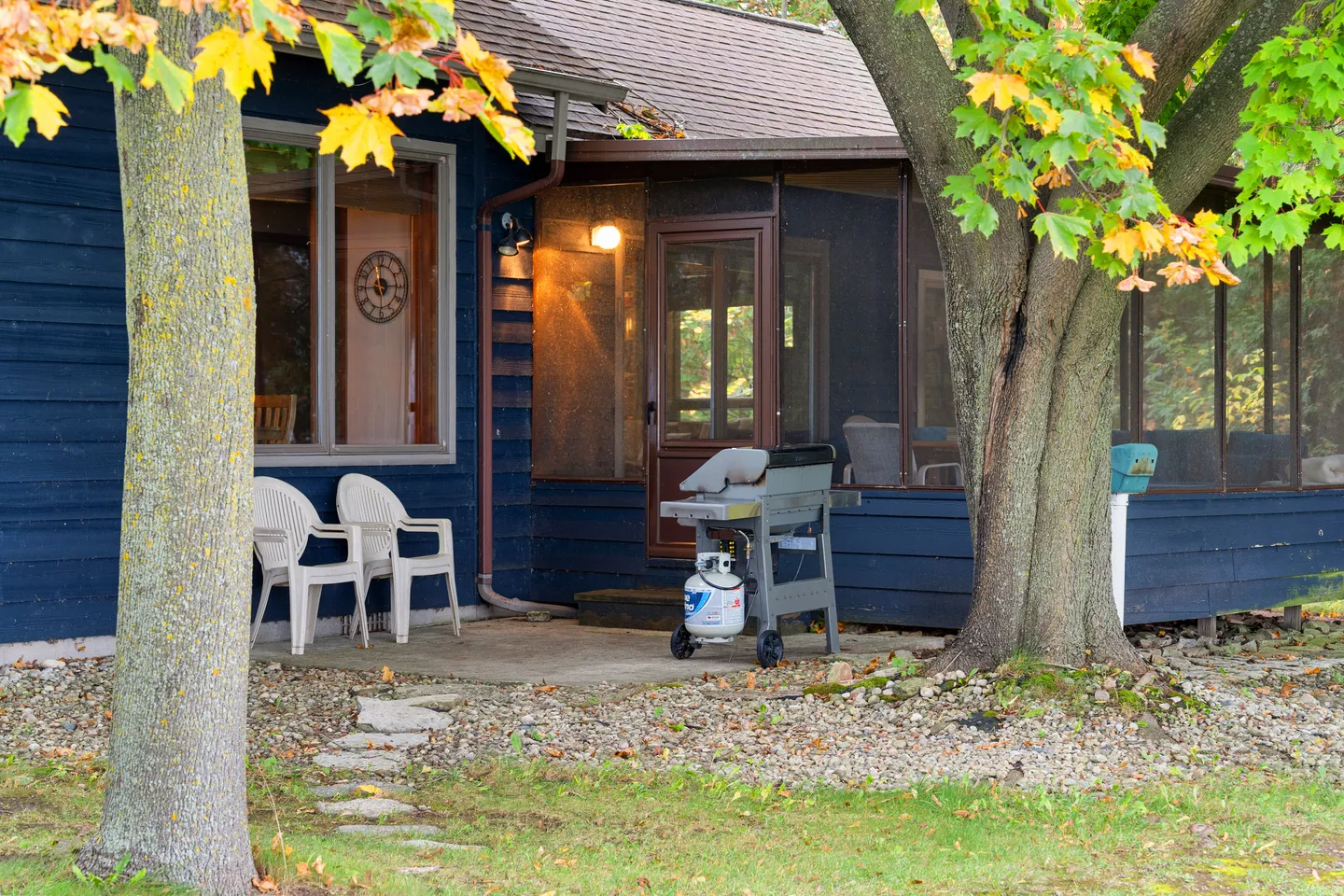 Exterior patio with white plastic chairs and a barbecue grill next to a blue house with a screened porch.