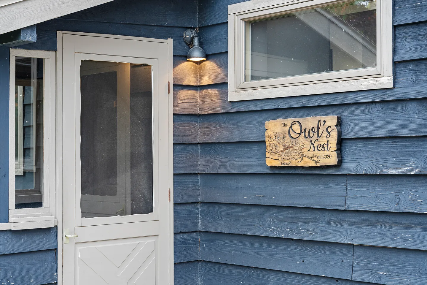 Exterior entrance featuring blue horizontal siding, a light-colored screen door, an illuminated sconce, and a wooden sign reading 'The Owl's Nest'.