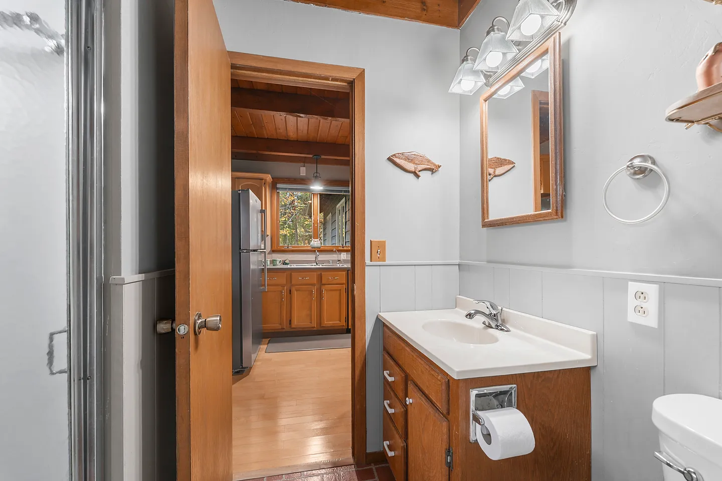 Bathroom with a wood vanity, mirror, and light fixture, looking through an open doorway into the kitchen.