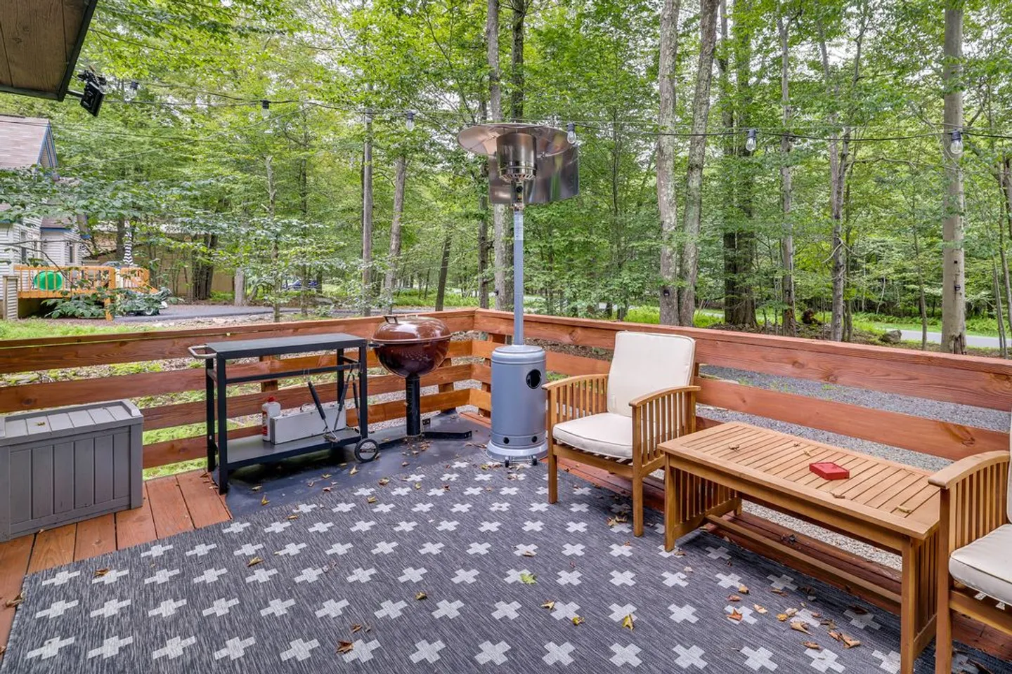 Outdoor wooden deck with an outdoor rug, patio heater, barbecue kettle grill, preparation cart, and wooden armchairs facing lush green woods.
