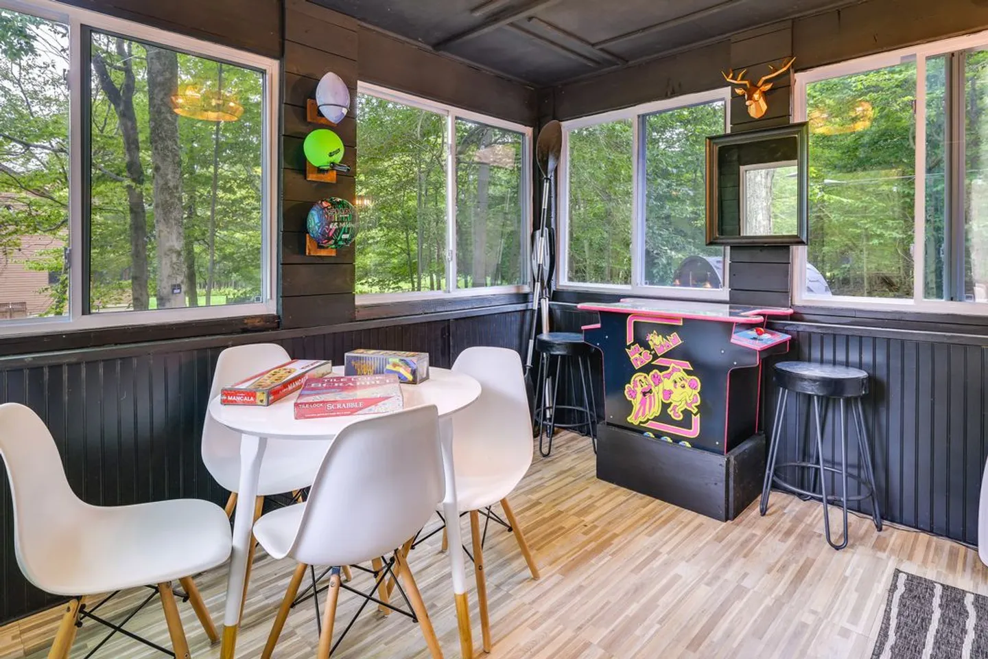 Sunroom game room with dark paneled walls, windows looking out to trees, a white table with board games and chairs, and an arcade console with barstools.