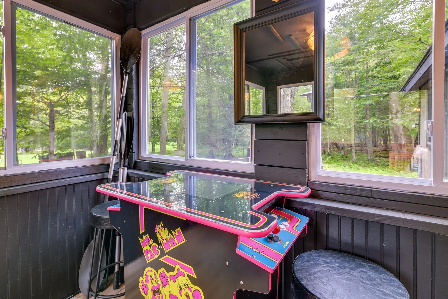 A Ms. Pac-Man cocktail arcade game table inside a sunroom with large windows overlooking outdoor woods.
