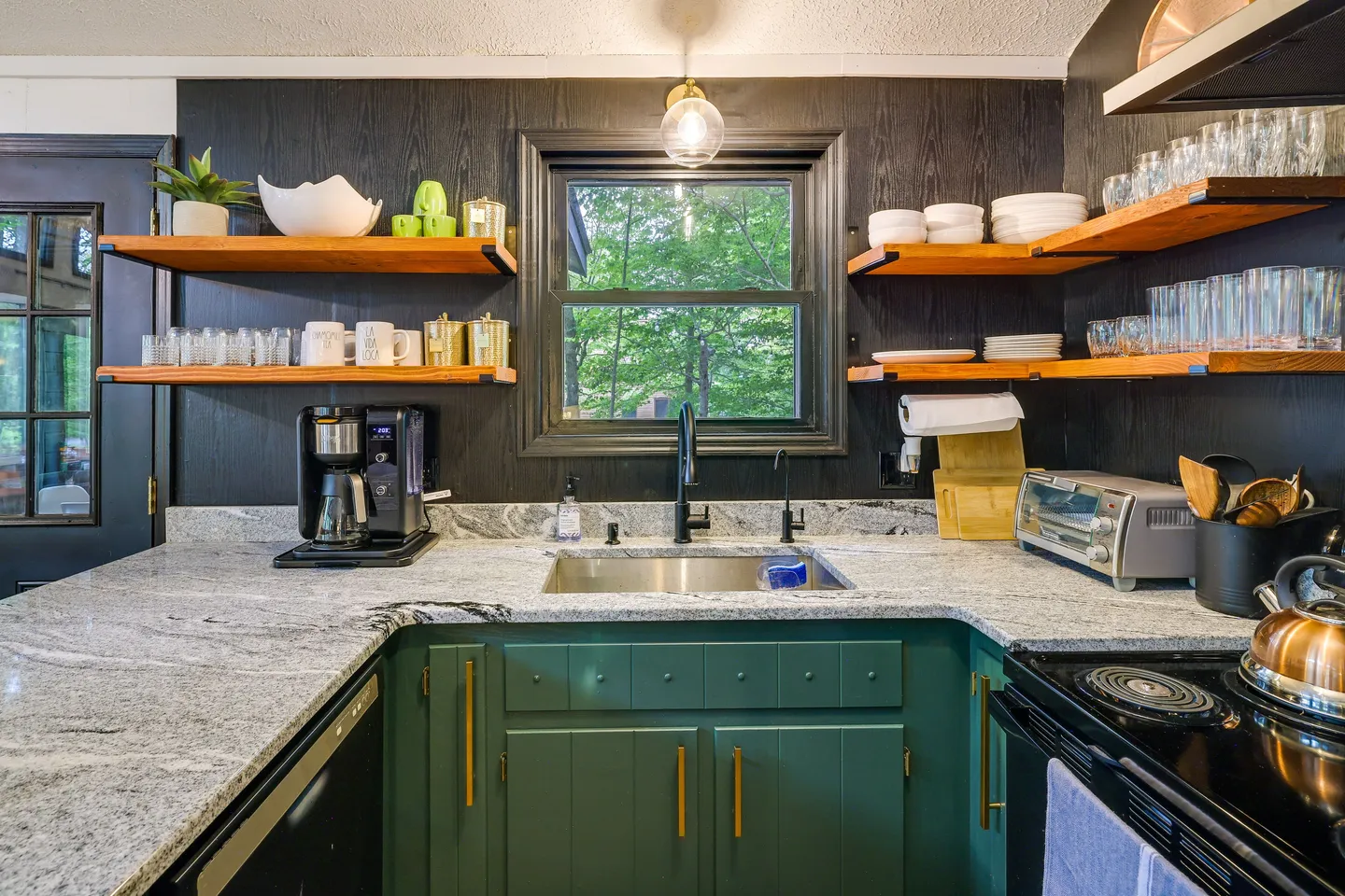 Kitchen sink area featuring dark green lower cabinets, granite countertops, and open wooden shelving against a black wood-paneled wall.