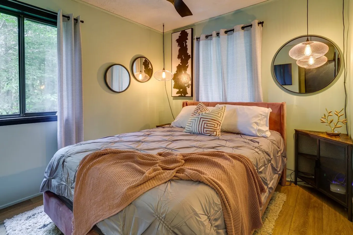 Bedroom with a made bed, light green walls, circular mirrors, hanging pendant lights, and two curtained windows.