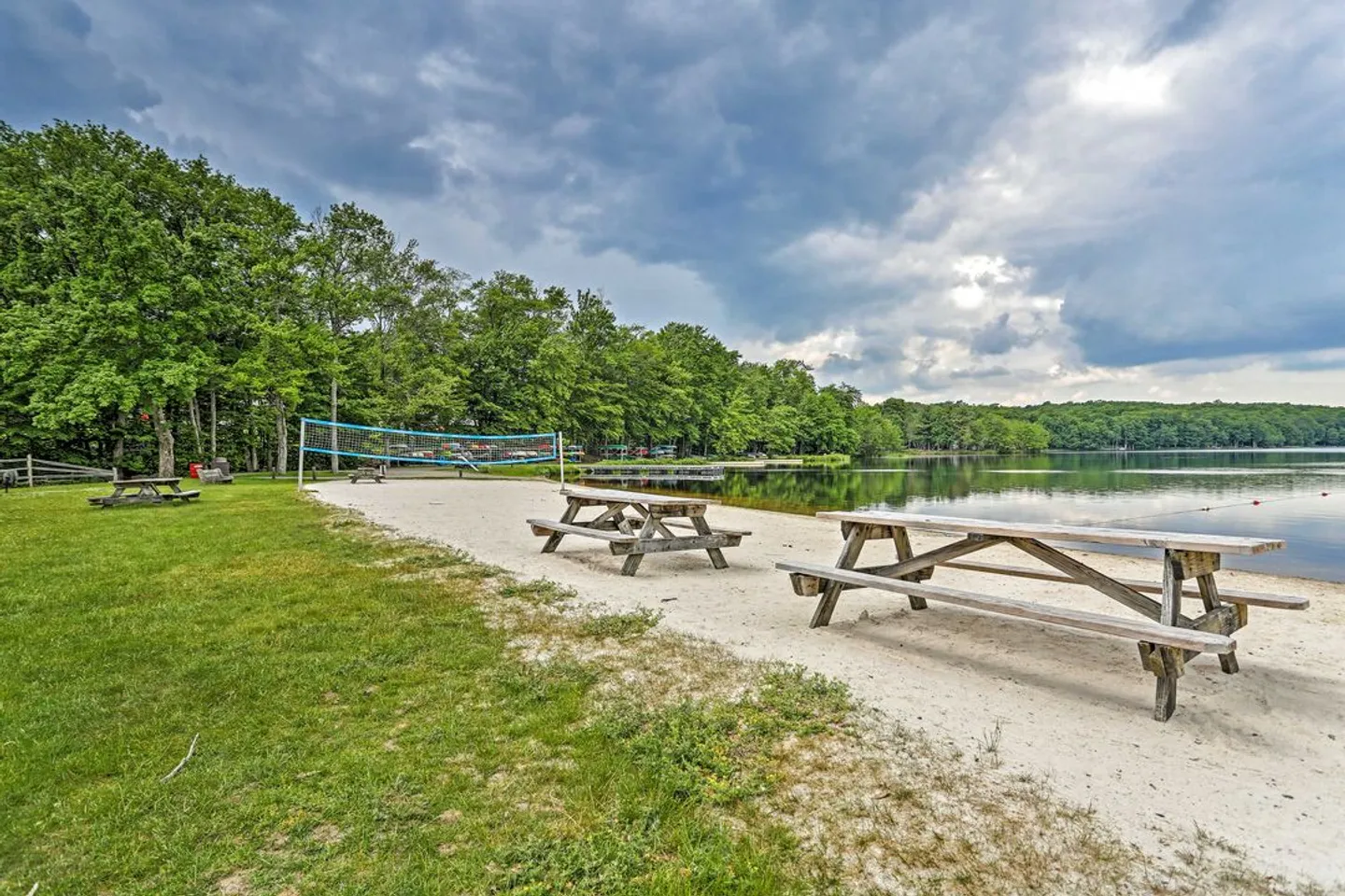 A sandy beach area along a lake with wooden picnic tables, a volleyball net, and surrounding trees under a cloudy sky.