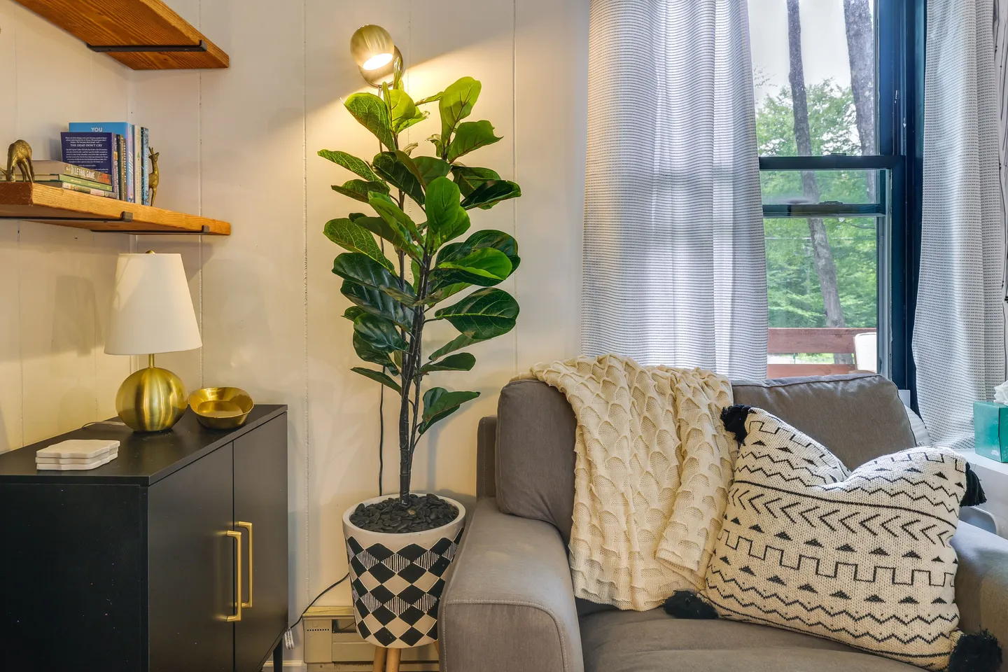 Cozy reading nook featuring an armchair with throw blanket and pillow, potted indoor plant, side cabinet with lamp, and window with curtains.