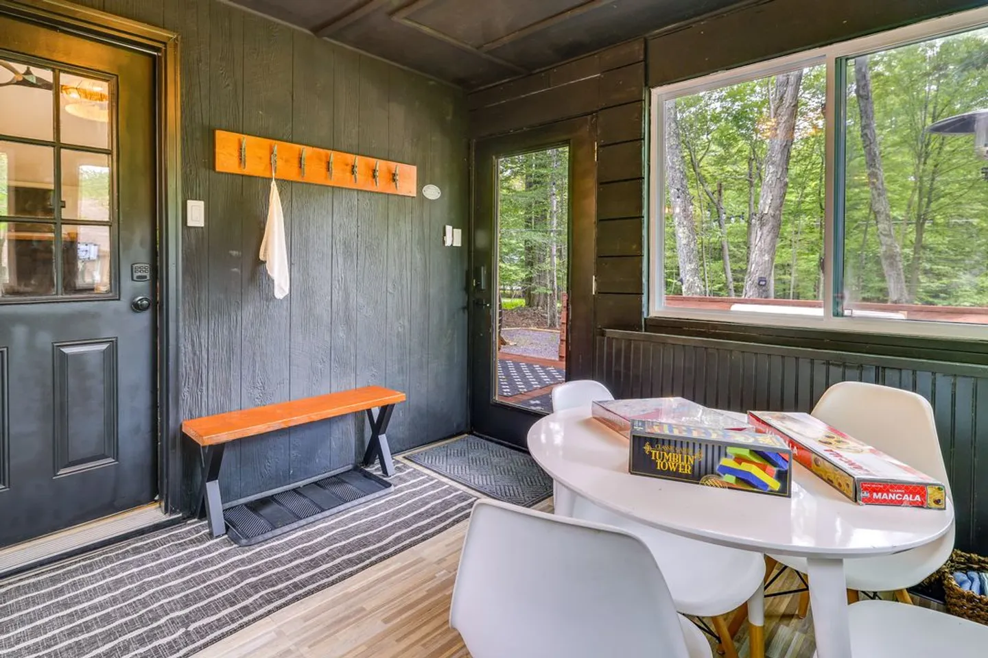 Sunroom entryway with dark paneled walls, a small table with chairs and board games, a wooden bench, and large windows looking outside to the woods.