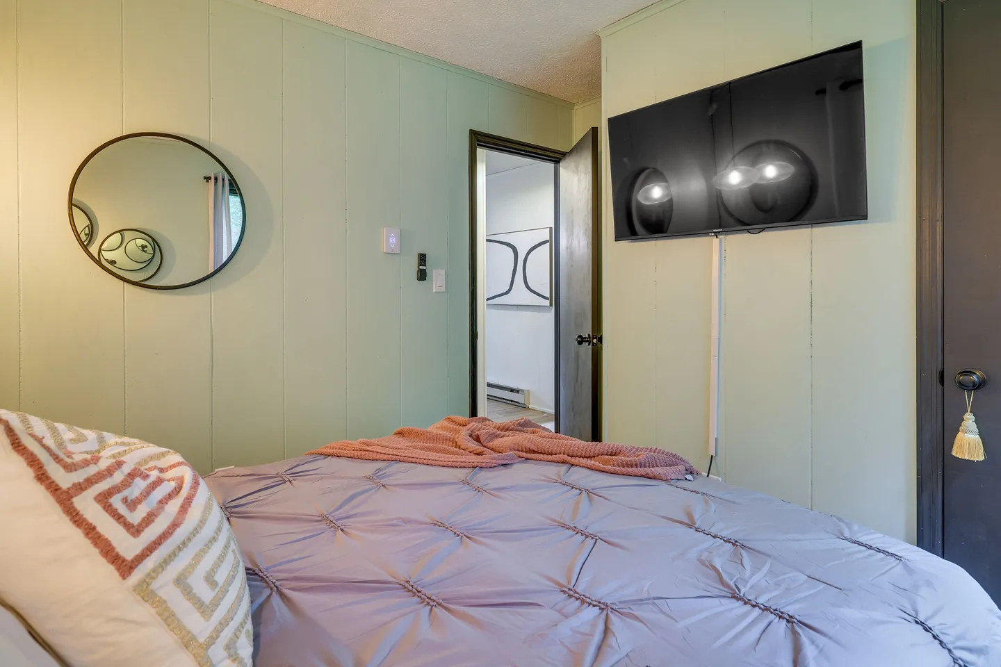 Bedroom with light green paneled walls, a mounted television, a circular mirror, and a made bed in the foreground.