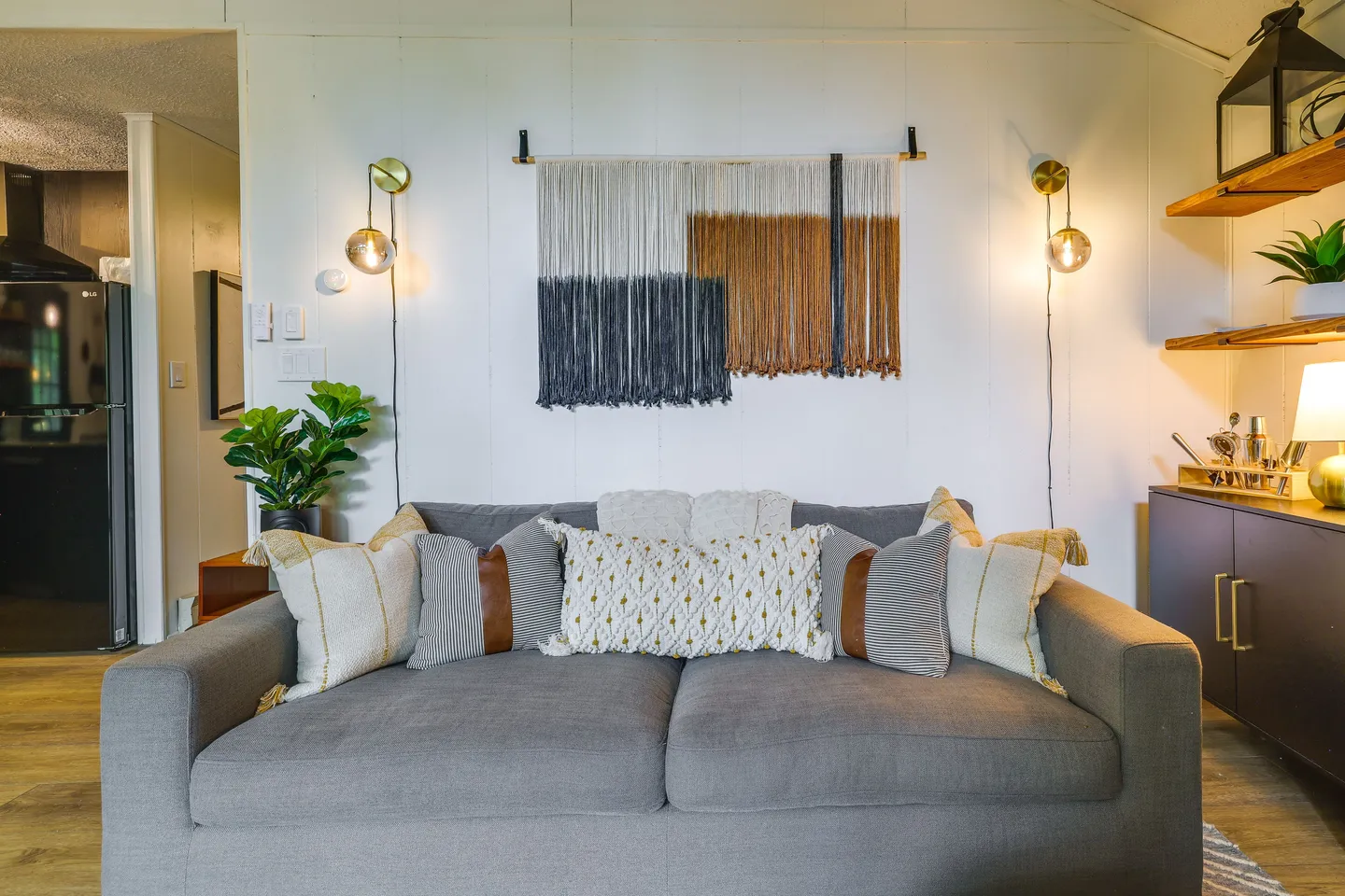 A gray upholstered sofa adorned with patterned throw pillows in a living room, positioned in front of a white wall featuring a yarn wall hanging and two mounted sconce lights.