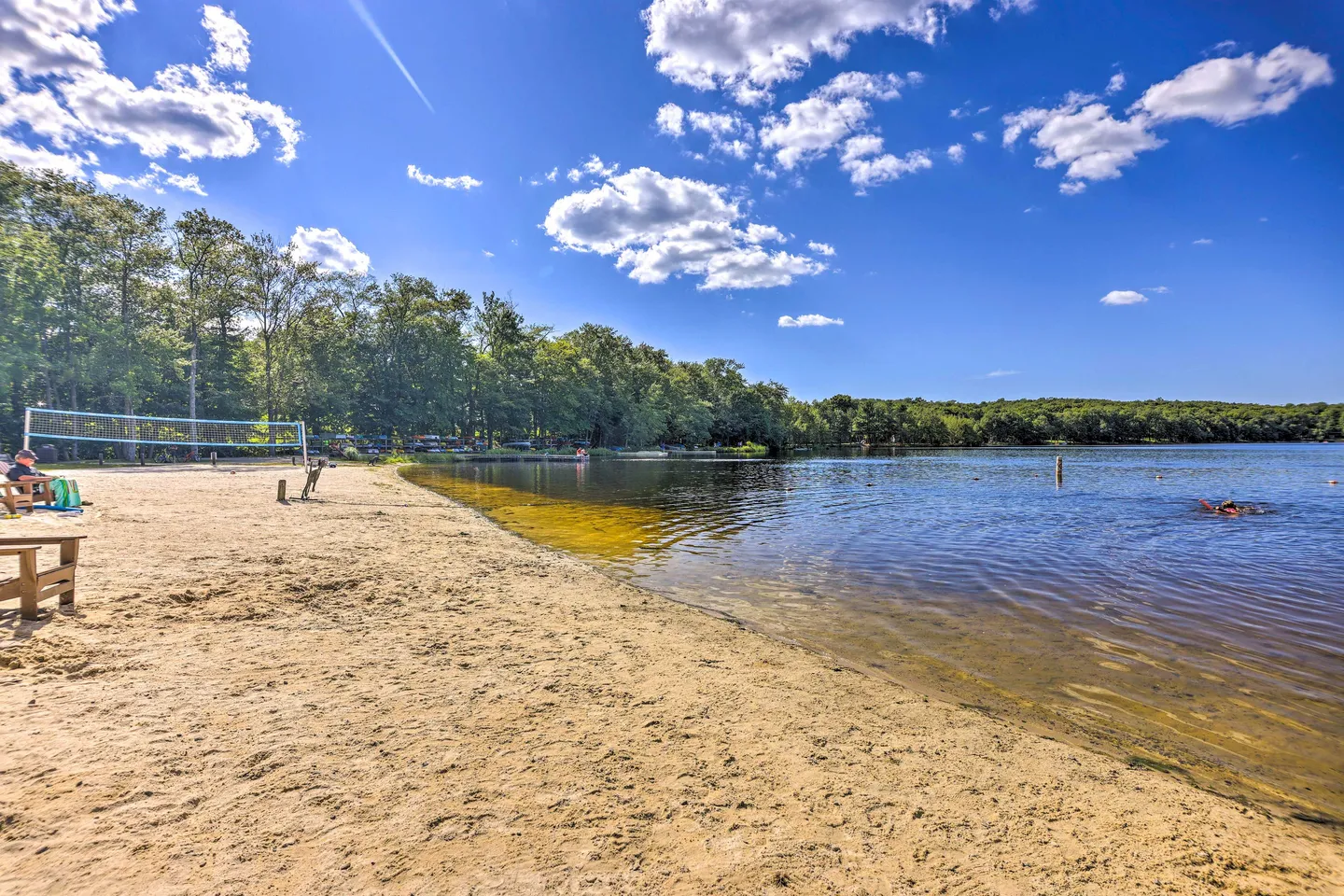 Sandy beach area bordering a lake, featuring a volleyball net and surrounded by dense green trees under a blue sky.