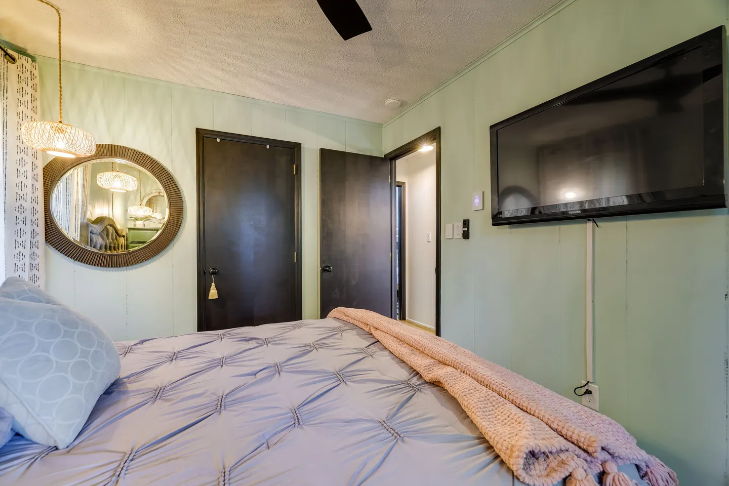 Bedroom with a bed in the foreground, light green paneled walls, a wall-mounted television, and dark interior doors.