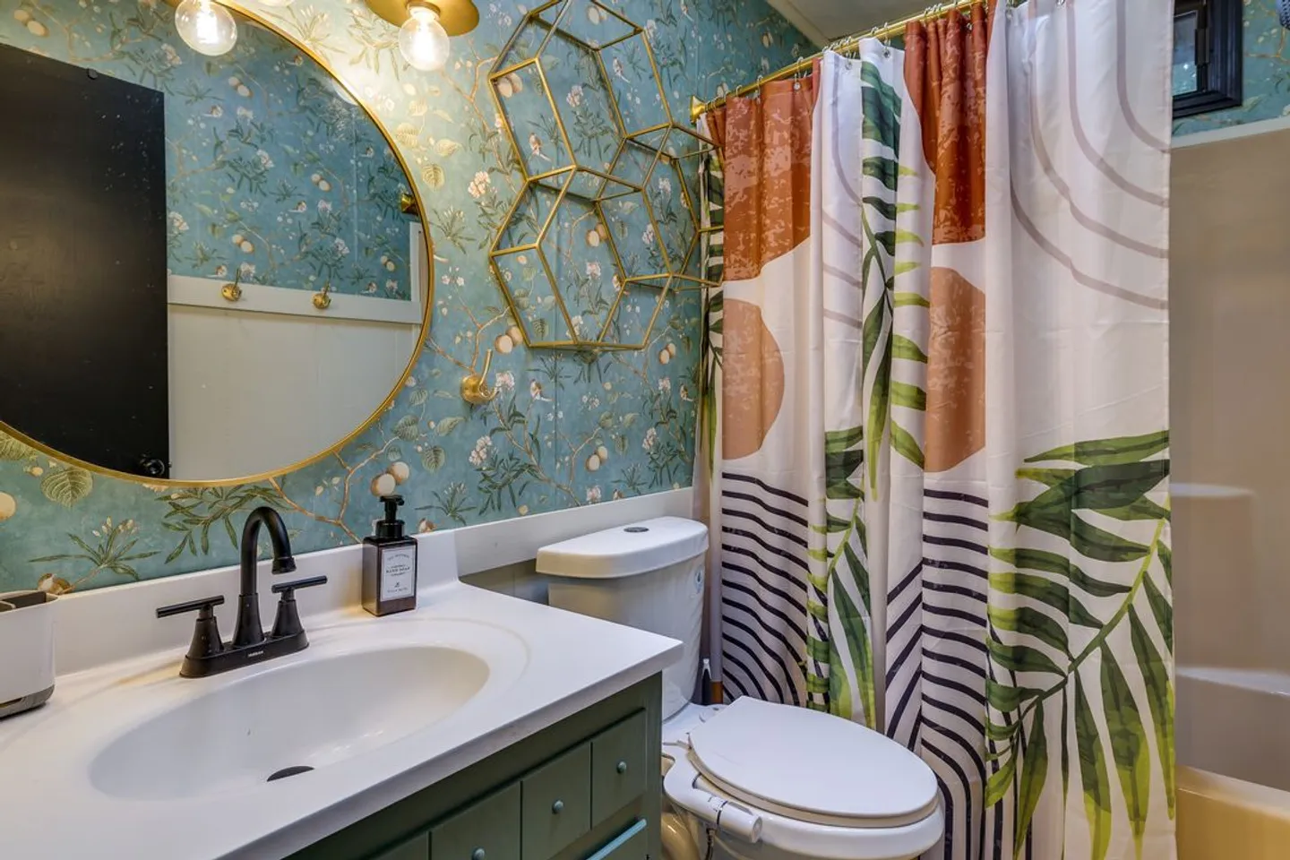 Bathroom with blue patterned wallpaper, a round mirror, vanity sink, toilet, and patterned shower curtain.