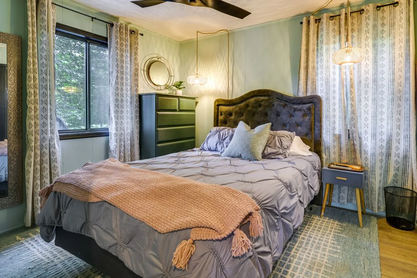 Bedroom with a tufted headboard, patterned rug, tall green dresser, and windows with patterned curtains.