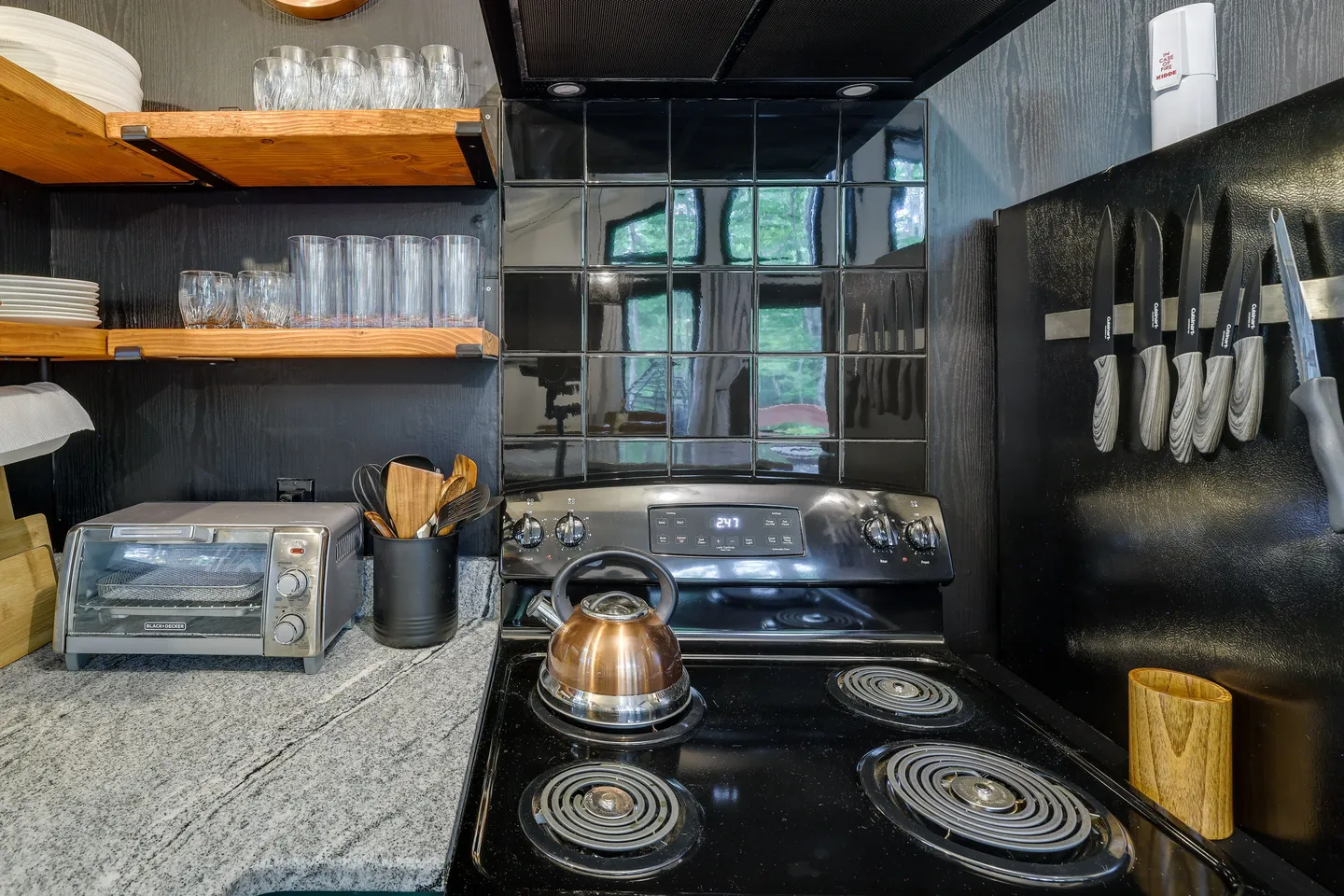 Kitchen counter area with an electric stove, copper kettle, toaster oven, and open shelving holding glassware.
