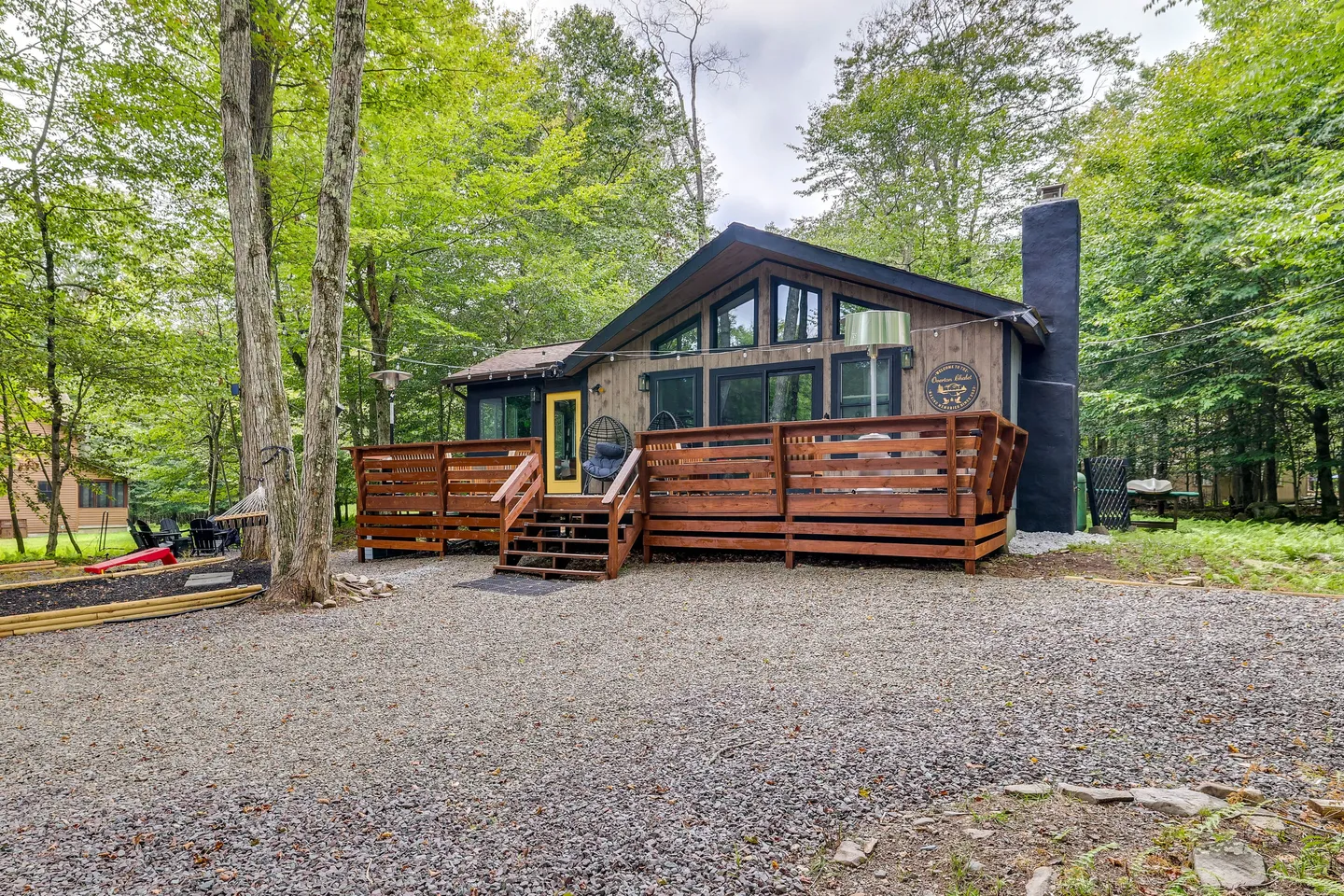 Exterior view of a cabin with a large front wooden deck, bright yellow front door, and gravel driveway surrounded by trees.
