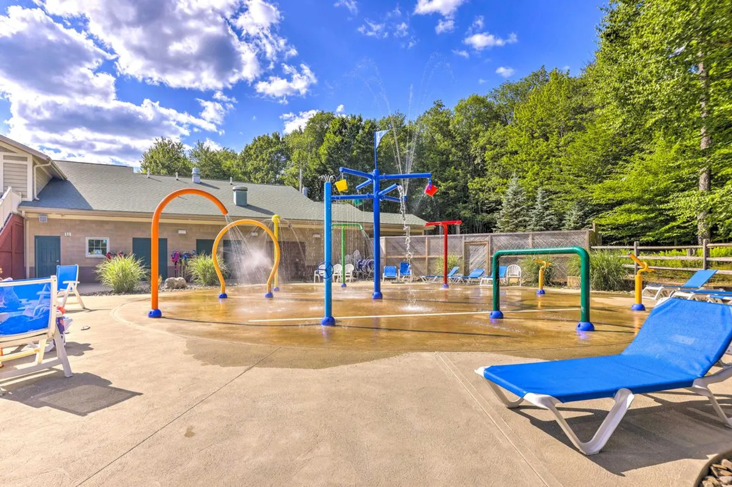 Outdoor community splash pad with colorful spraying water features and surrounding lounge chairs under a blue sky.