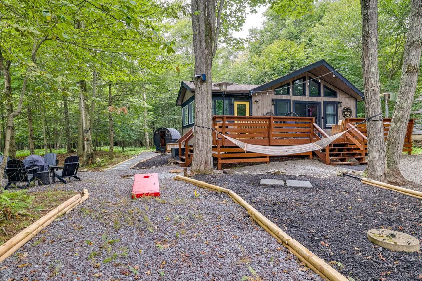 Exterior backyard view of a wood-sided cabin in the woods, featuring an elevated deck, hammock, fire pit with chairs, and lawn games.