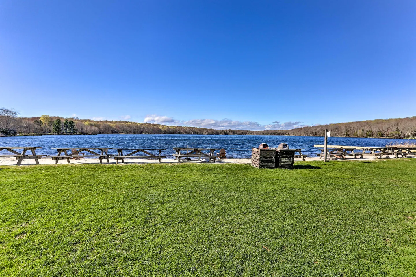 A wide view of a lakefront beach area with picnic tables, trash receptacles, and a grassy lawn under a clear blue sky.