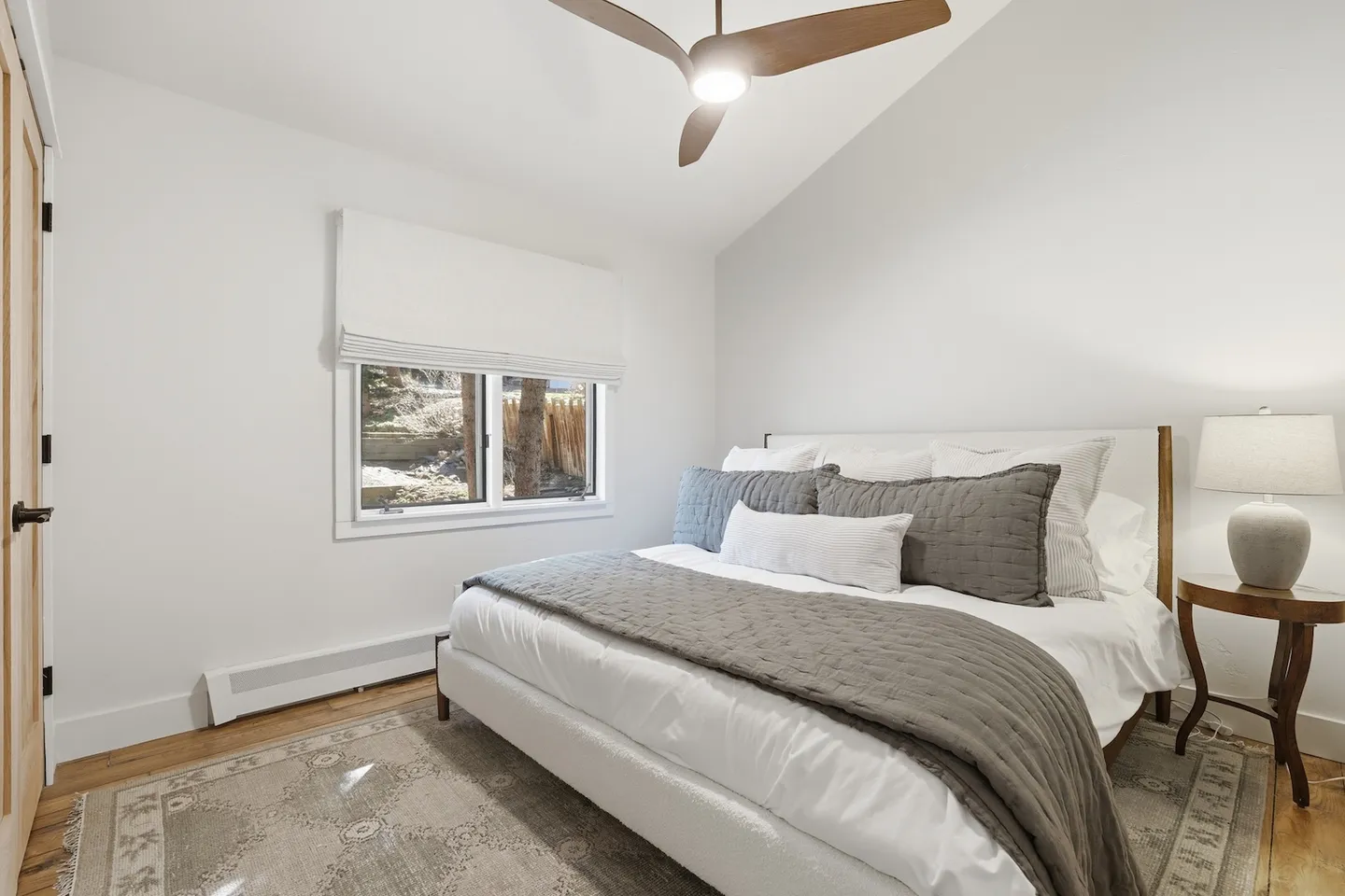 Bedroom with a vaulted ceiling, wood-blade ceiling fan, white upholstered bed, and patterned area rug over wood flooring.