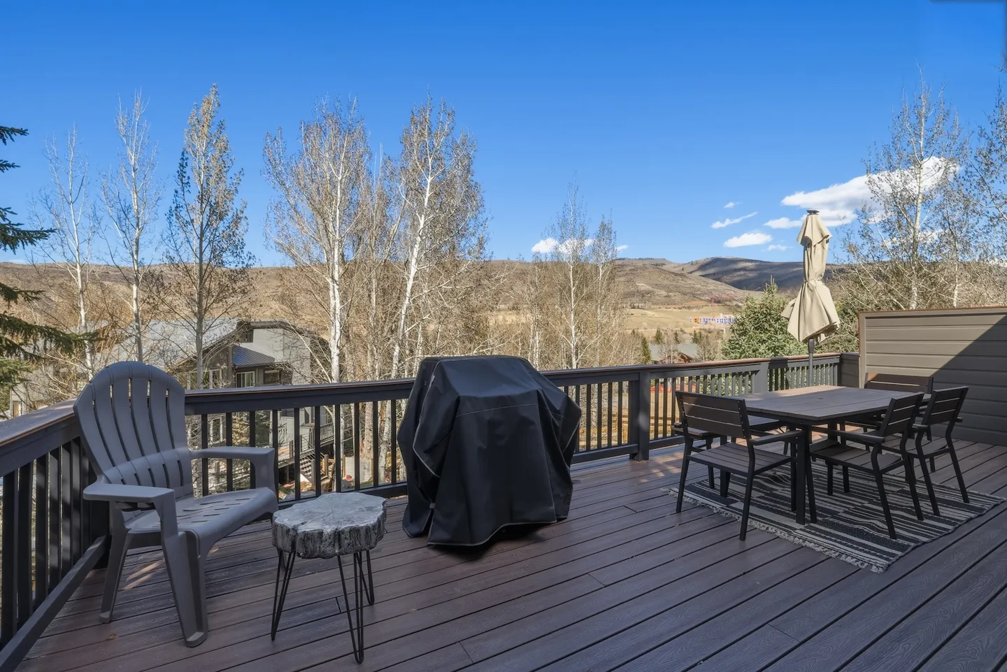 Outdoor deck with an Adirondack chair, small table, covered grill, and dining set overlooking hills and trees under a clear blue sky.