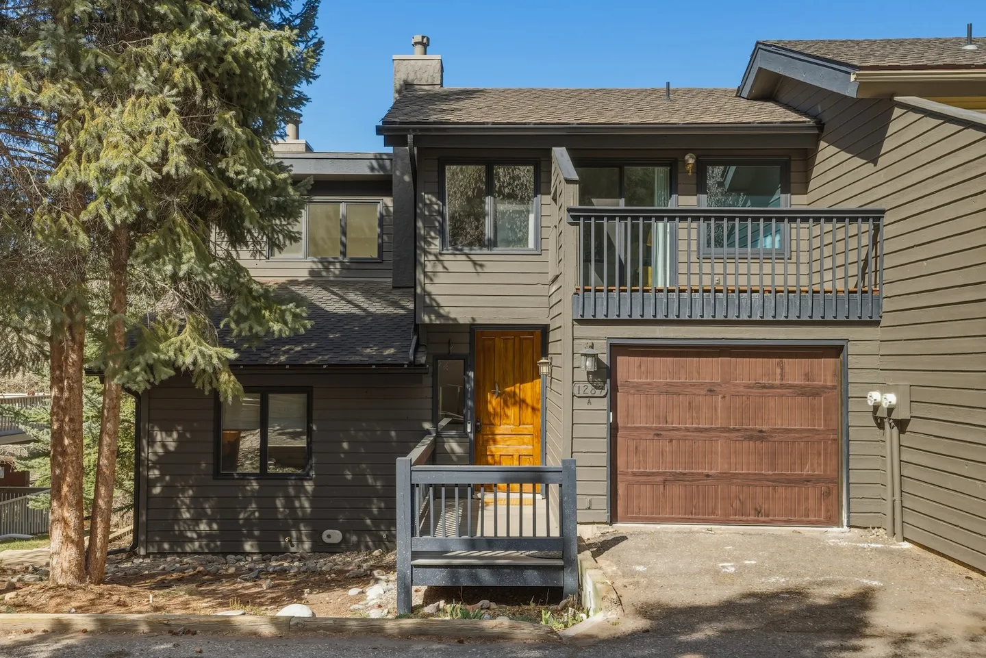 Exterior of a two-story multi-level home with dark wood siding, a wooden front door, an attached single-car garage, and an upper balcony.