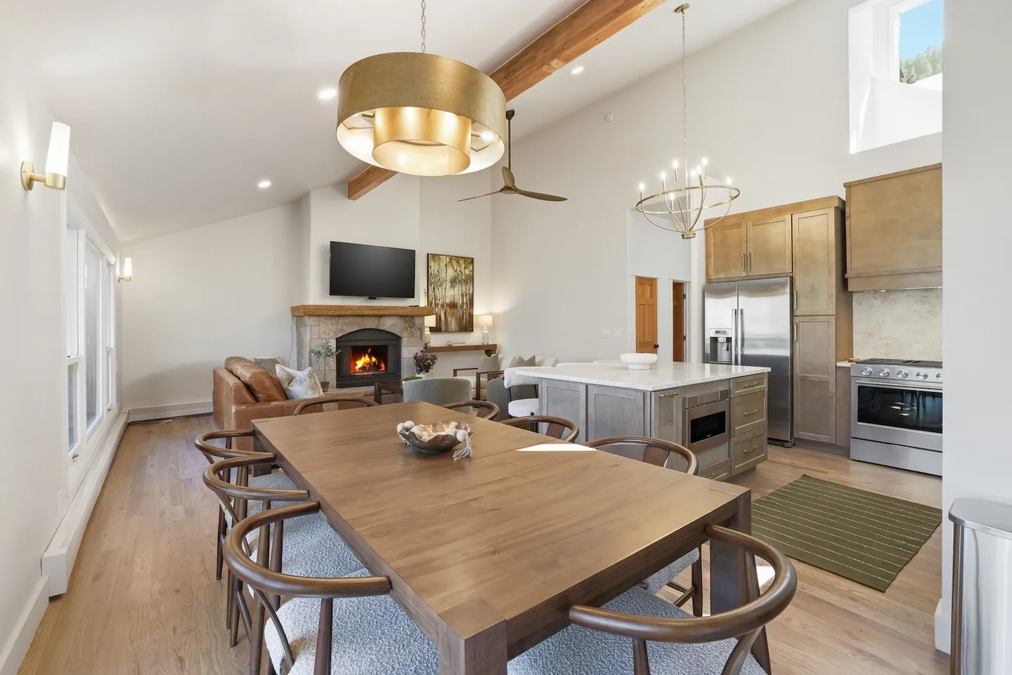 Open-concept dining, kitchen, and living area with vaulted ceilings, a wooden dining table, kitchen island, and a stone fireplace.