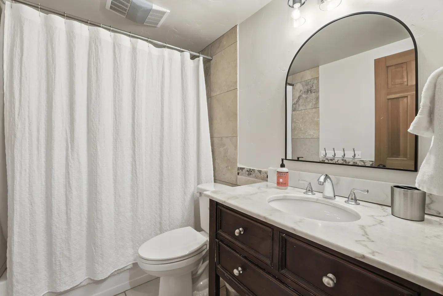Bathroom featuring a dark wood vanity with a marble-style countertop, arched mirror, toilet, and shower with a white curtain.