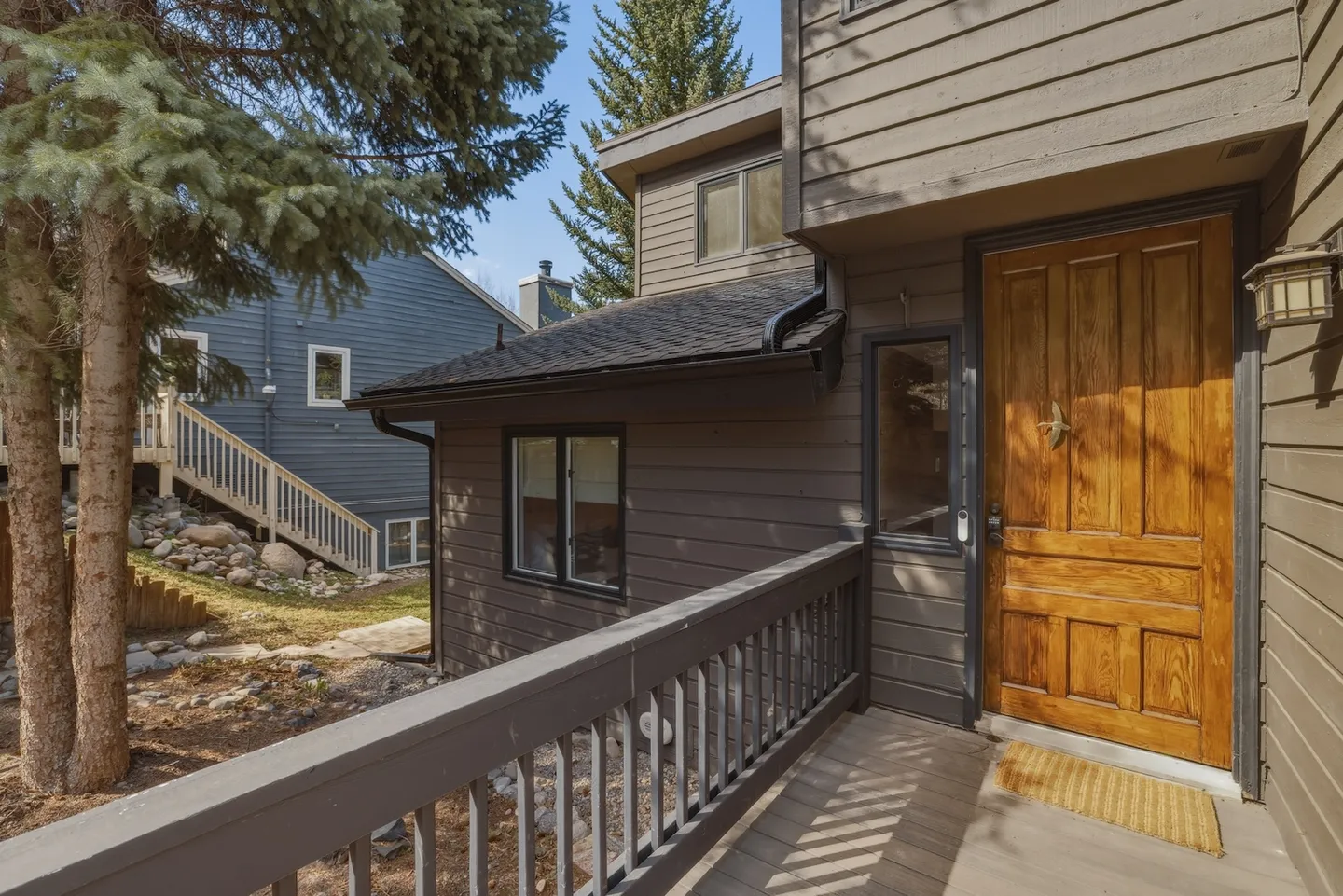 Exterior entryway of a home featuring a wooden front door, small porch with railing, brown siding, and nearby evergreen trees under daylight.